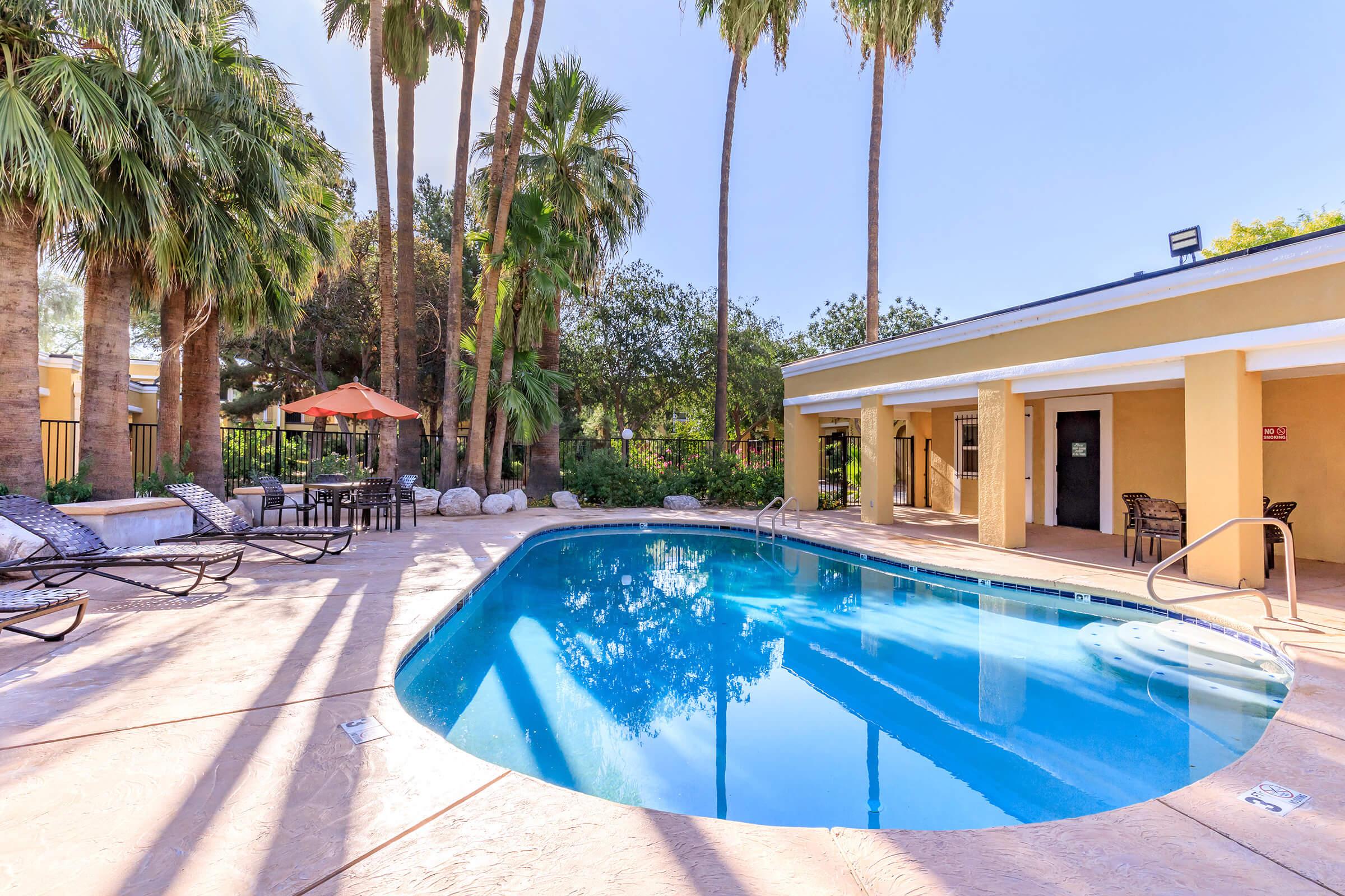 Clear blue swimming pool surrounded by tall palm trees and lounge chairs. An orange umbrella provides shade, and a building with a yellow exterior is visible in the background. Sunlight casts shadows on the pool deck, creating a serene and inviting atmosphere.