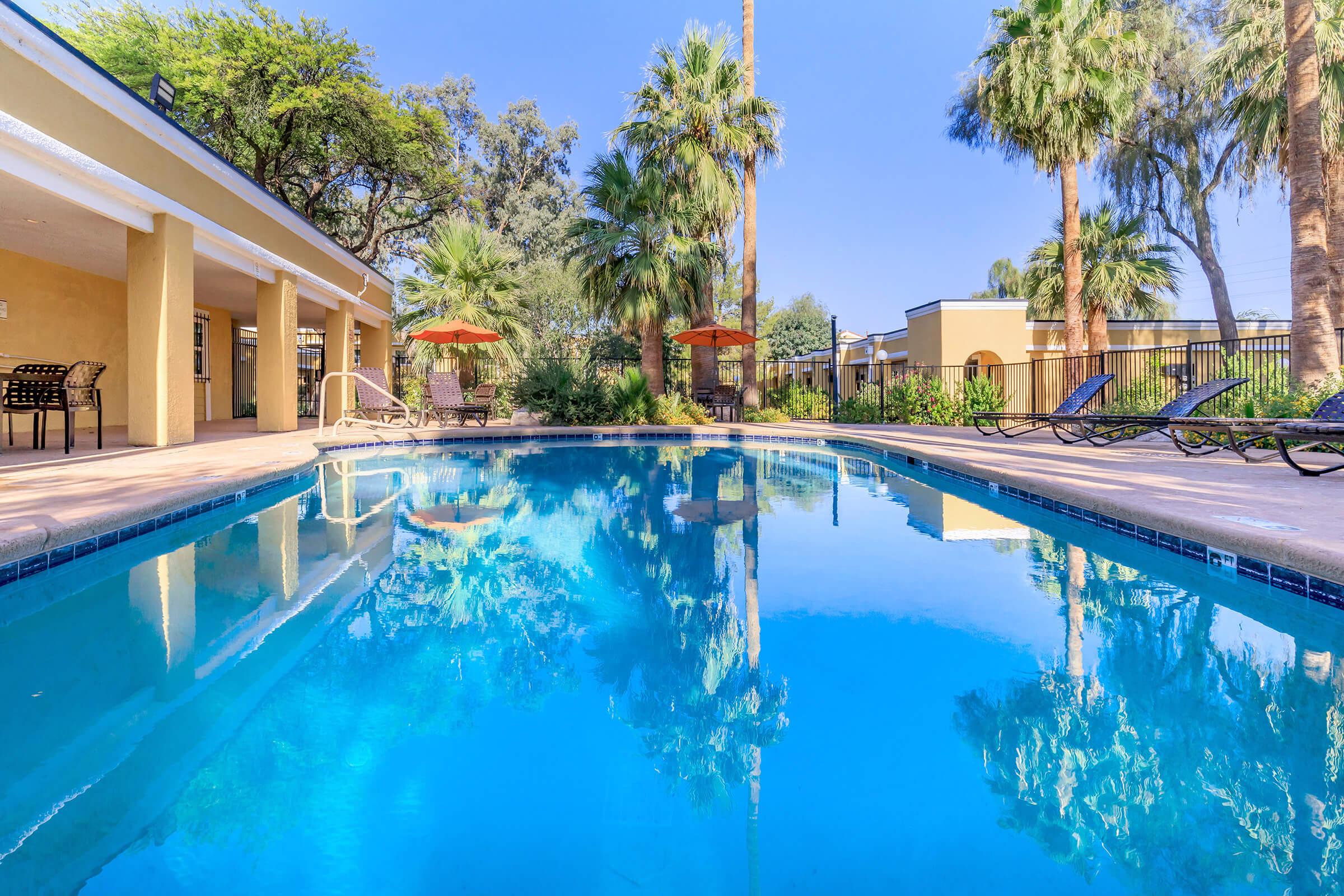 A serene pool area surrounded by palm trees, featuring lounge chairs and orange umbrellas. The clear blue water reflects the bright sky and lush greenery. Comfortable seating is available alongside the pool, creating an inviting atmosphere for relaxation.