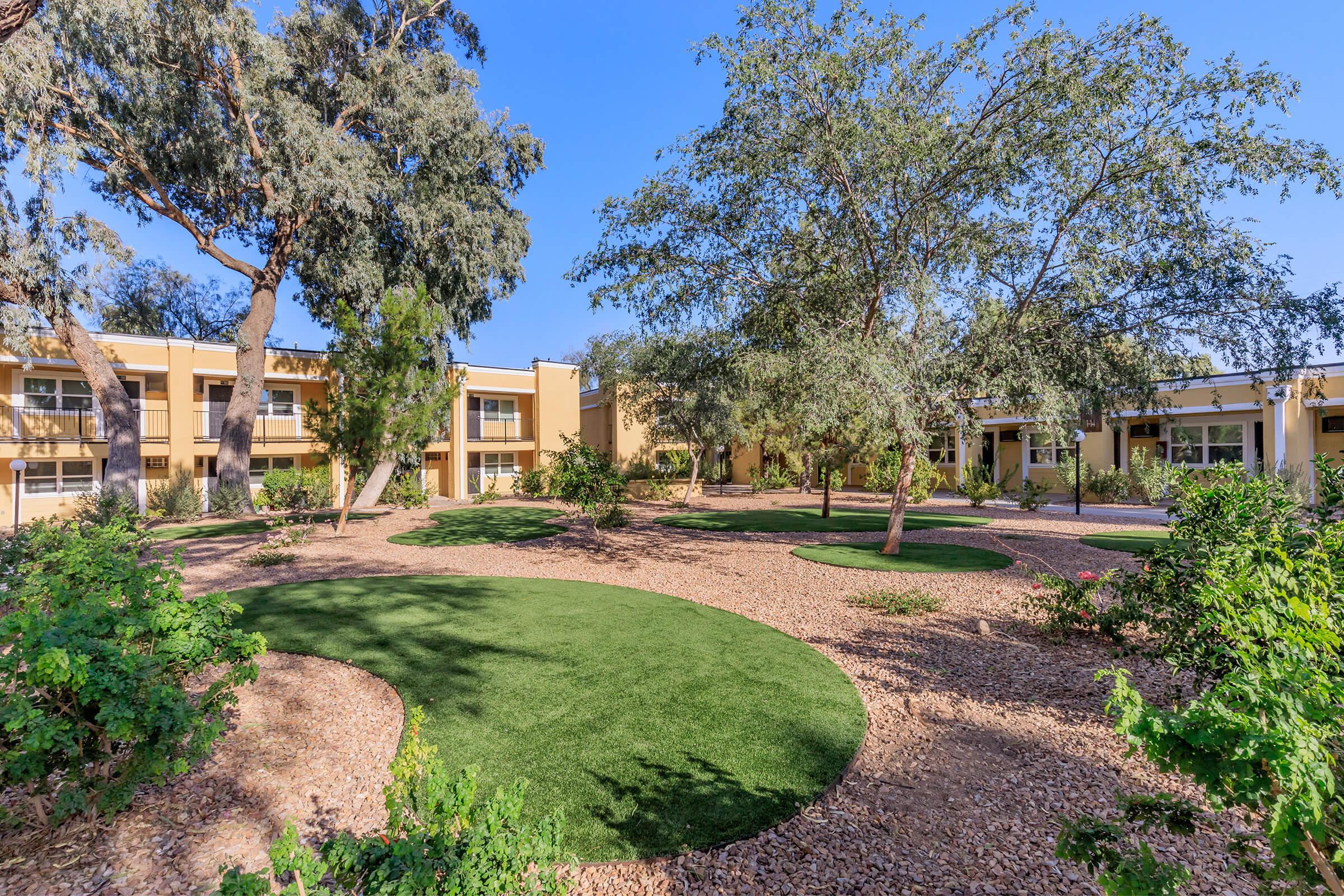 A peaceful courtyard featuring manicured green grass and landscaped areas surrounded by trees and shrubbery. Two-story buildings with balconies are visible in the background, under a clear blue sky. The setting is calm and inviting, ideal for relaxation and outdoor activities.
