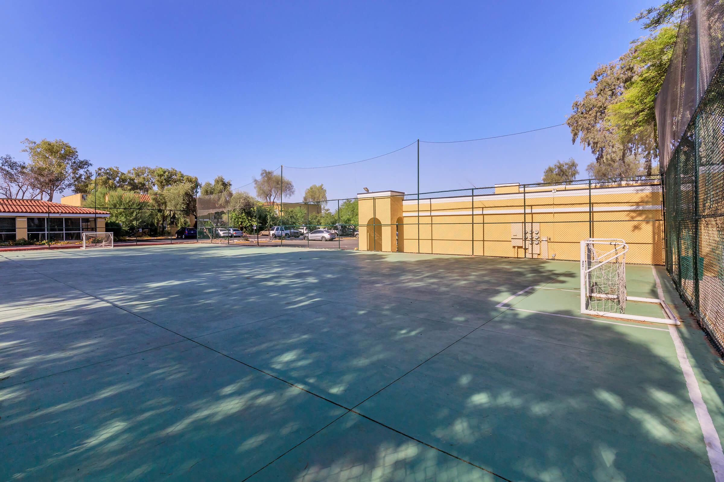 Empty outdoor sports court with a green surface, a soccer goal on the left, and a fenced area in the background. Trees are visible around the court, and a small building is partially seen on the left side. The sky is clear and blue.