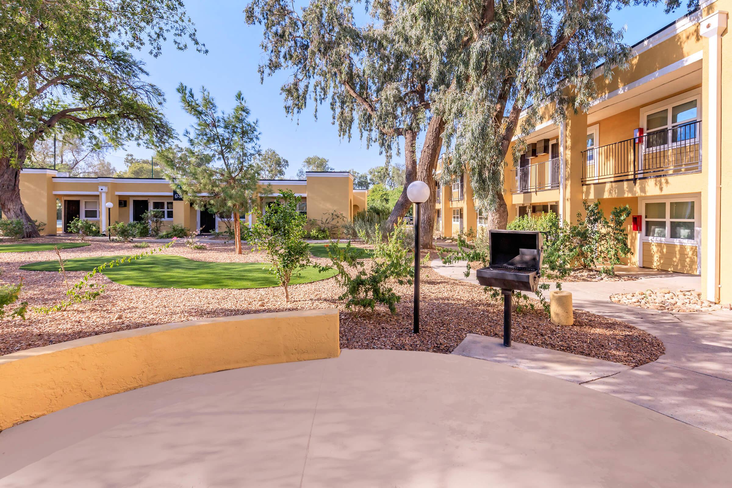 A landscaped courtyard with greenery, trees, and a barbecue grill. Surrounding the area are two-story buildings with yellow walls and balconies, featuring pathways and well-maintained grounds. The setting appears inviting and peaceful, perfect for outdoor gatherings.