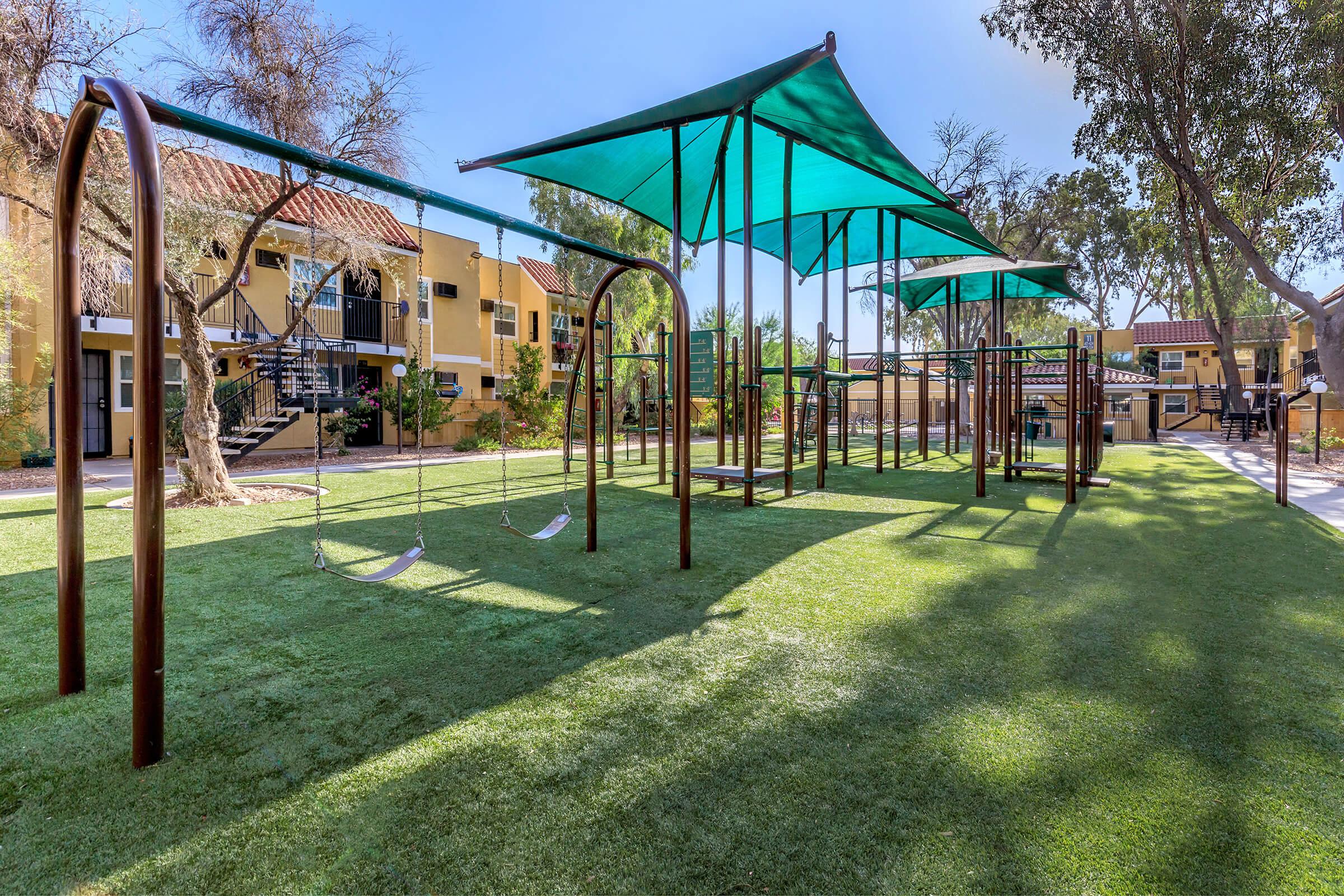 A playground area featuring two swings under shaded structures, surrounded by green grass and trees, with residential buildings visible in the background. The scene is bright and inviting, ideal for children to play and enjoy the outdoors.