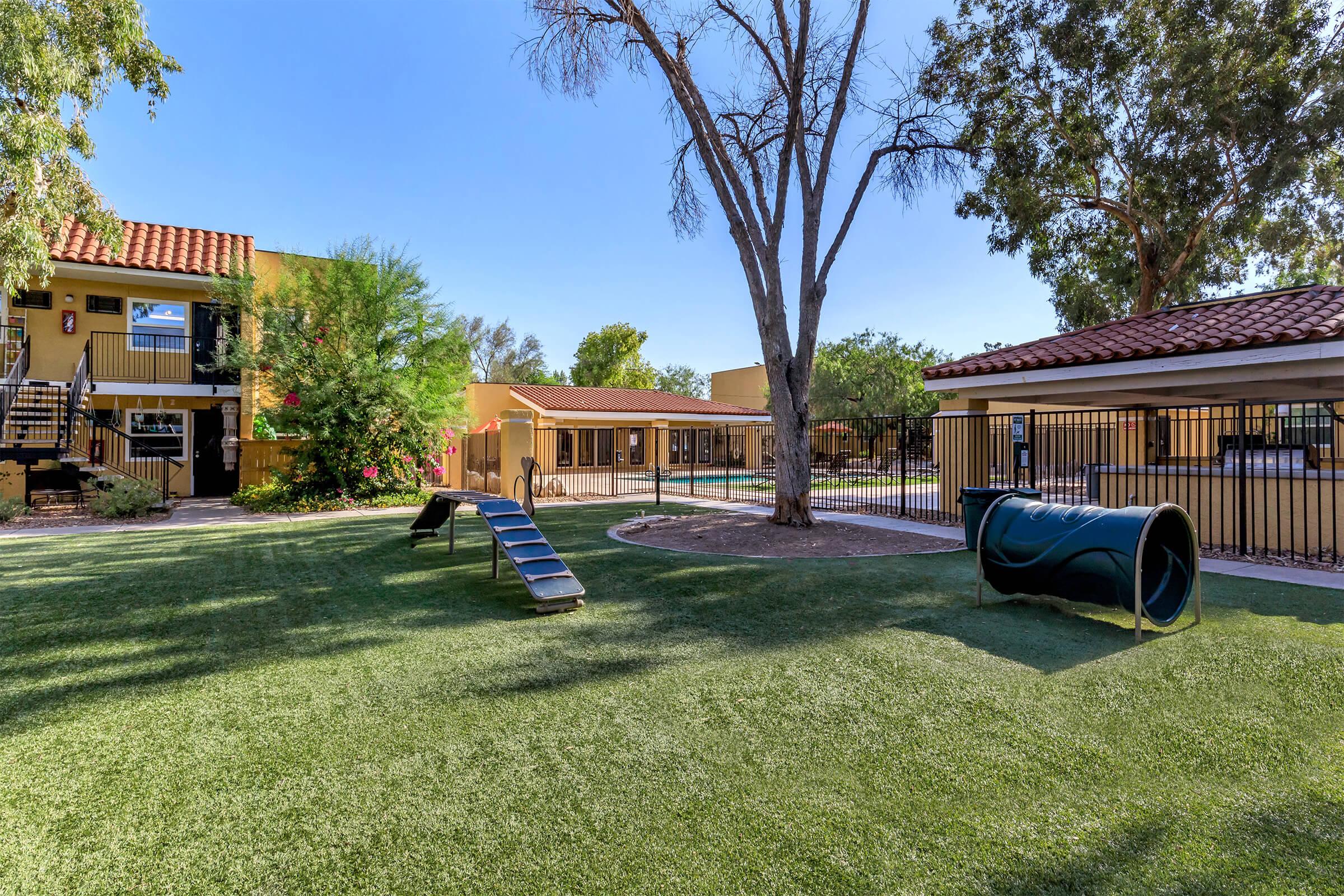 A landscaped outdoor area featuring artificial grass, a slide, and a tunnel for pets. Surrounding buildings are visible, along with a pool and trees providing shade. The space is fenced and designed for recreation and relaxation.