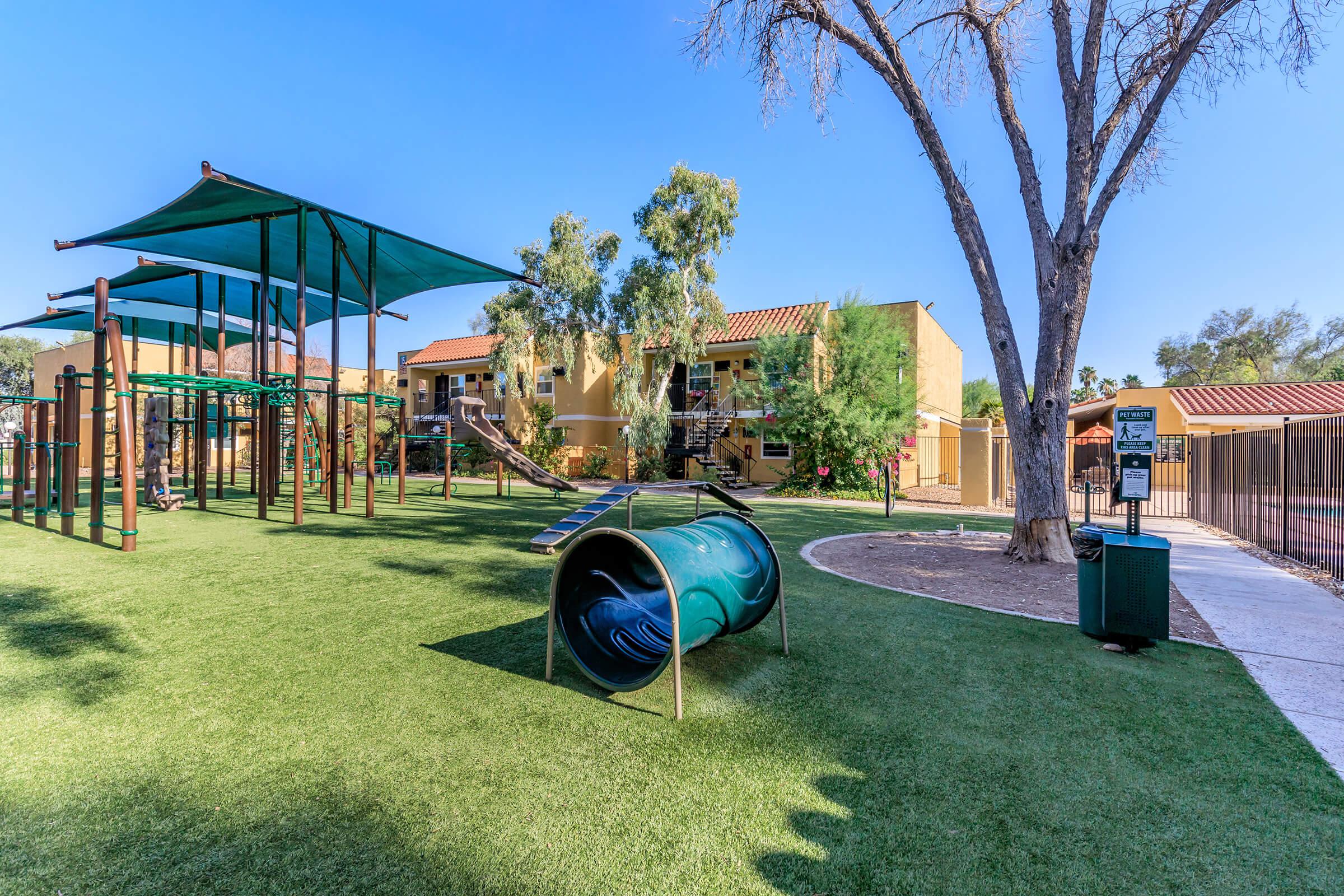 Playground area featuring green synthetic grass, climbing structures, and a tunnel slide. Shaded areas provided by blue canopies. In the background, residential buildings are visible with a clear blue sky overhead. A trash bin and pathway are also present, creating an inviting outdoor space for children.