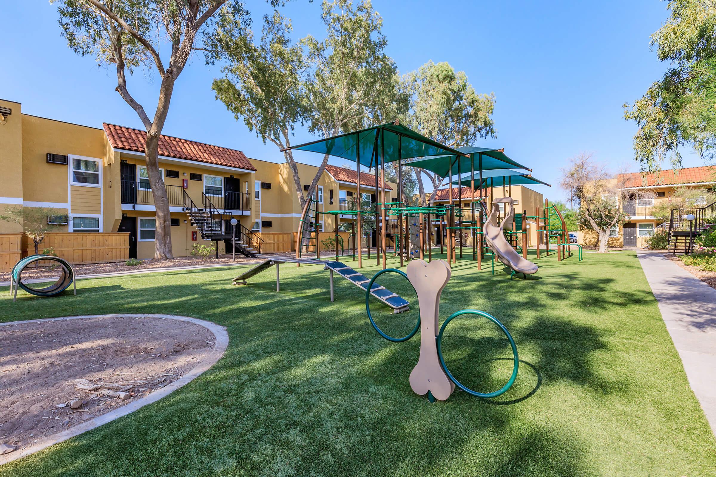 Playground area featuring a green grassy field with modern play equipment, including slides, climbing structures, and swings. Surrounding trees provide shade. Nearby are residential apartment buildings, contributing to a family-friendly atmosphere. Clear blue sky enhances the vibrant setting.