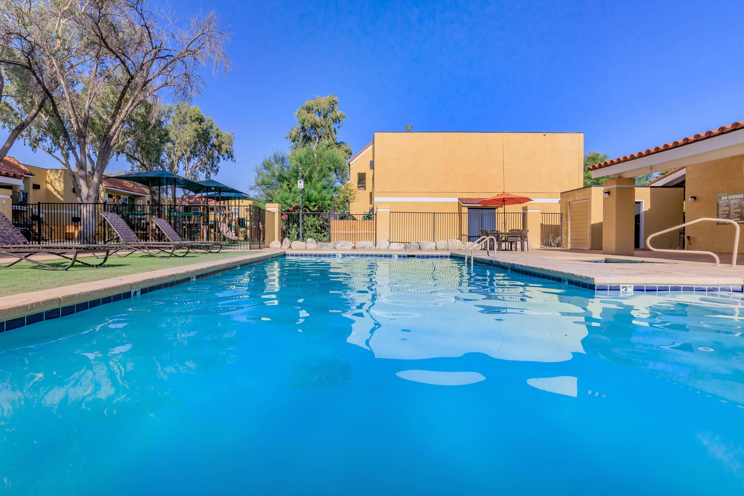 A clear blue swimming pool surrounded by lounge chairs and shaded areas. Buildings with yellow stucco walls are visible in the background, along with trees and outdoor seating. The sky is bright blue, indicating a sunny day.