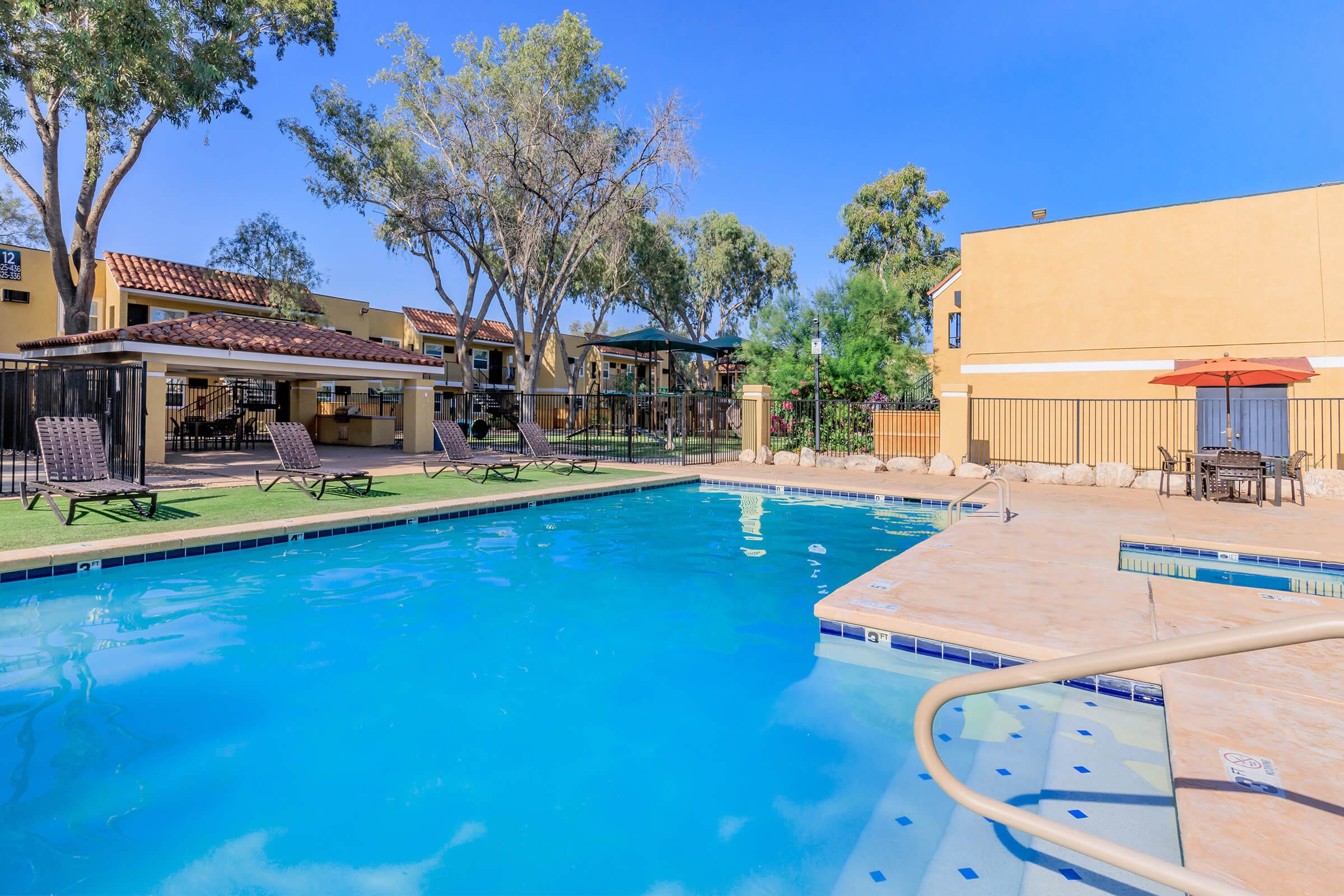 A serene pool area surrounded by lounge chairs and trees, with a clear blue sky overhead. In the background, there are buildings featuring a Mediterranean architectural style, and a patio area with an umbrella. The scene conveys a relaxing outdoor space ideal for leisure and swimming.