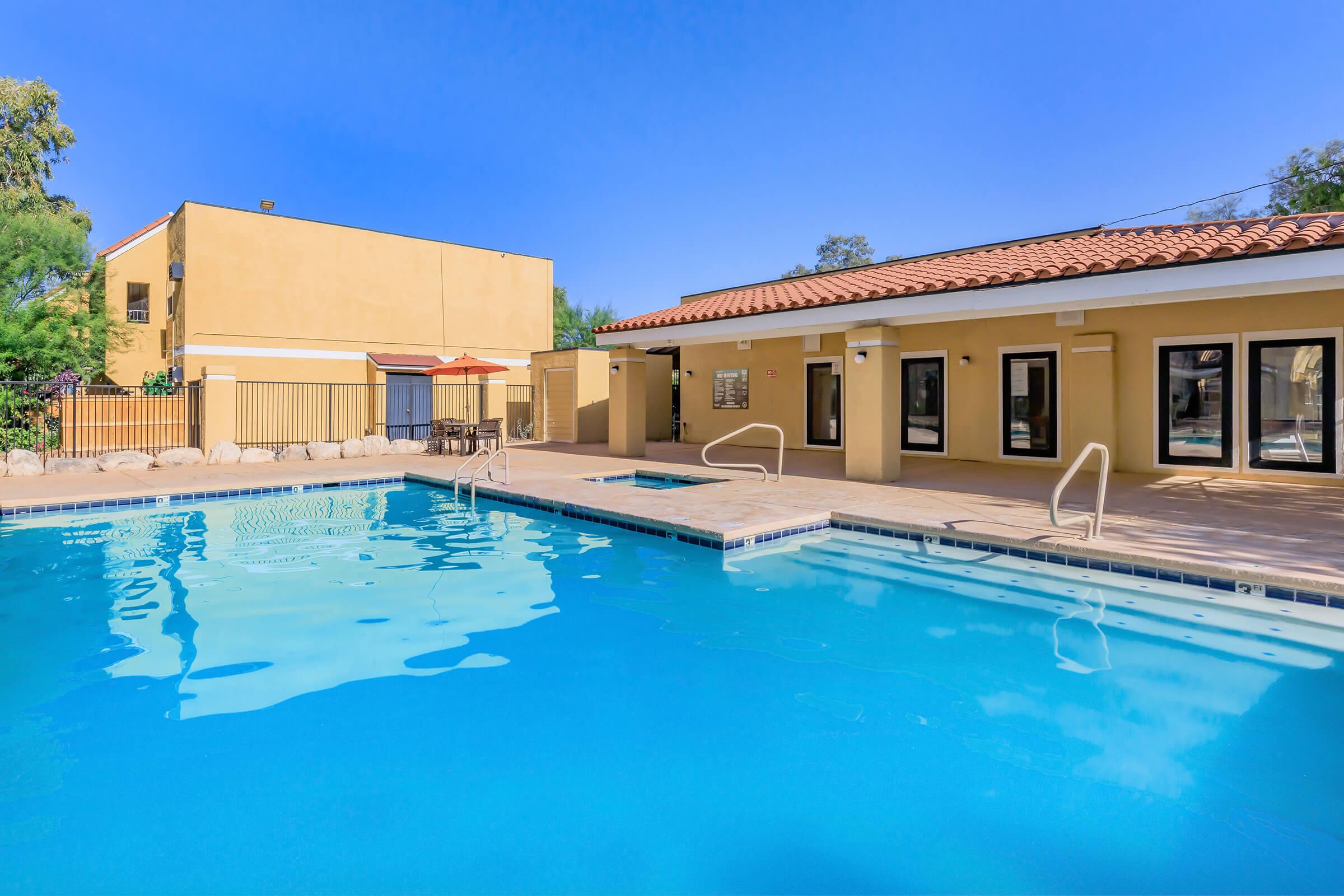 A clear blue swimming pool surrounded by a tiled area, with a building featuring a reddish-tiled roof in the background. There are lounge chairs and an umbrella nearby, under a bright blue sky. The scene is inviting and ideal for relaxation.