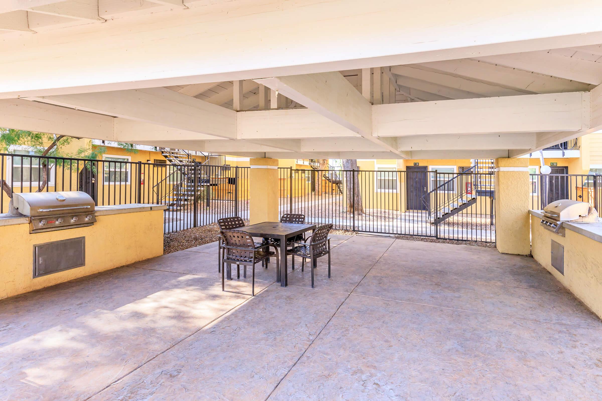 A covered outdoor seating area featuring a table and chairs, with two built-in grills on either side. The space is enclosed by a fence and has a concrete floor, surrounded by yellow walls and trees, providing a shaded area for dining or gatherings.