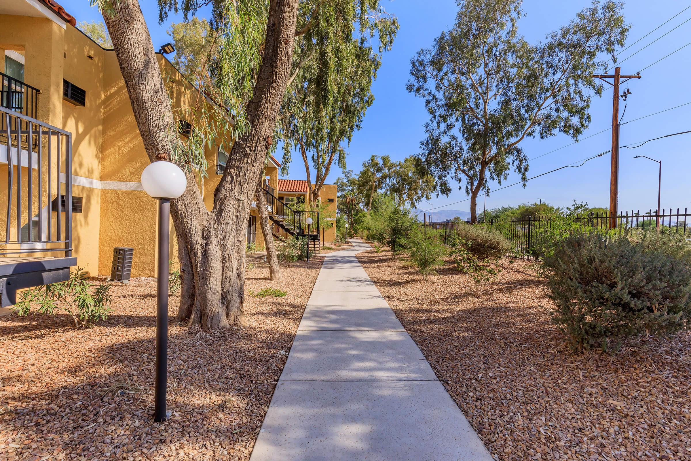 A walkway lined with trees and shrubs, leading through a landscaped area. Soft sunlight filters through the foliage, casting gentle shadows on the concrete path. In the background, buildings can be seen, adding to the serene outdoor atmosphere.