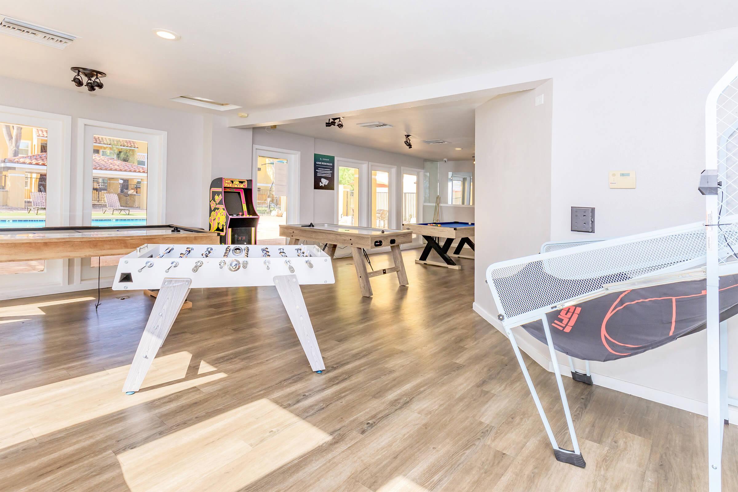Interior view of a recreational room featuring several game tables, including foosball and air hockey. The space has large windows allowing natural light in, with a classic arcade machine visible in the corner. Light wood flooring adds to the inviting atmosphere of the room.