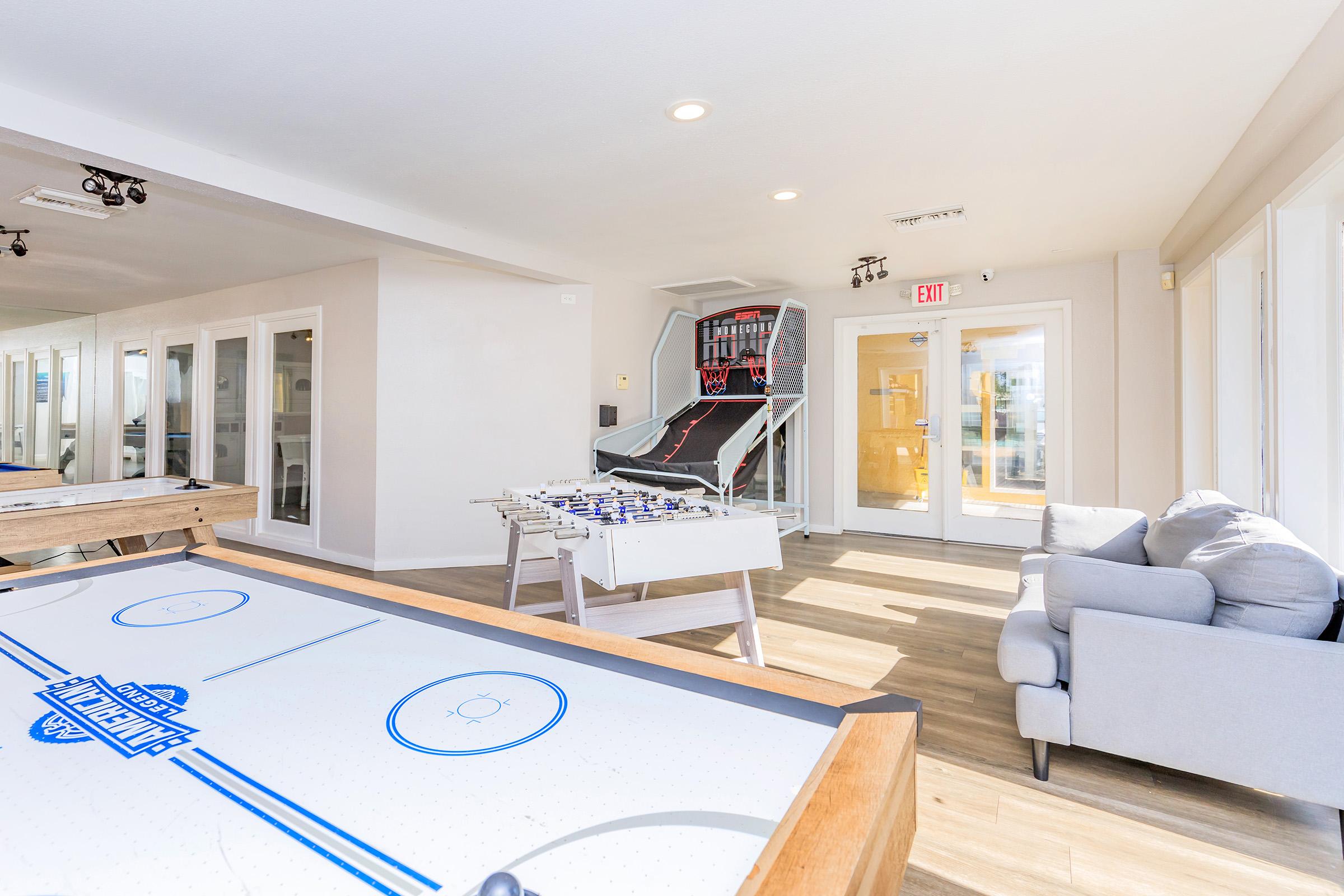 A bright recreational room featuring a foosball table, an air hockey table, and a seating area with a couch. Large windows provide natural light, and doors lead to an outdoor area. The modern design creates a welcoming atmosphere for leisure activities.