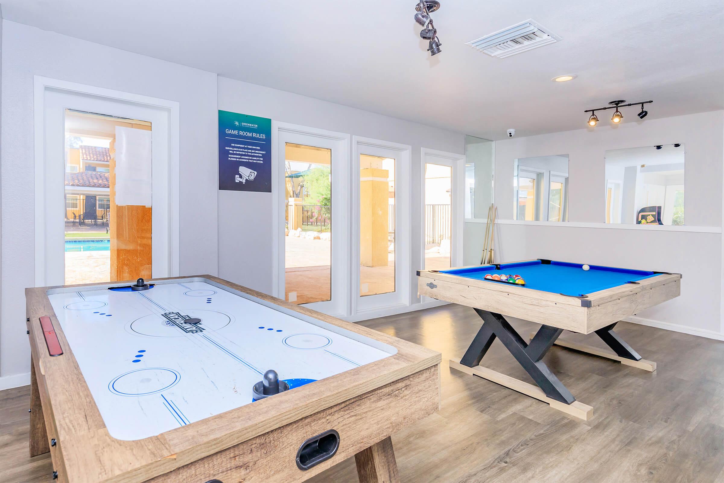 A game room featuring an air hockey table and a pool table. Large windows provide natural light and views of an outdoor pool area. The walls are painted in soft colors, and there is modern lighting overhead. A wall display includes information about game rules or features.
