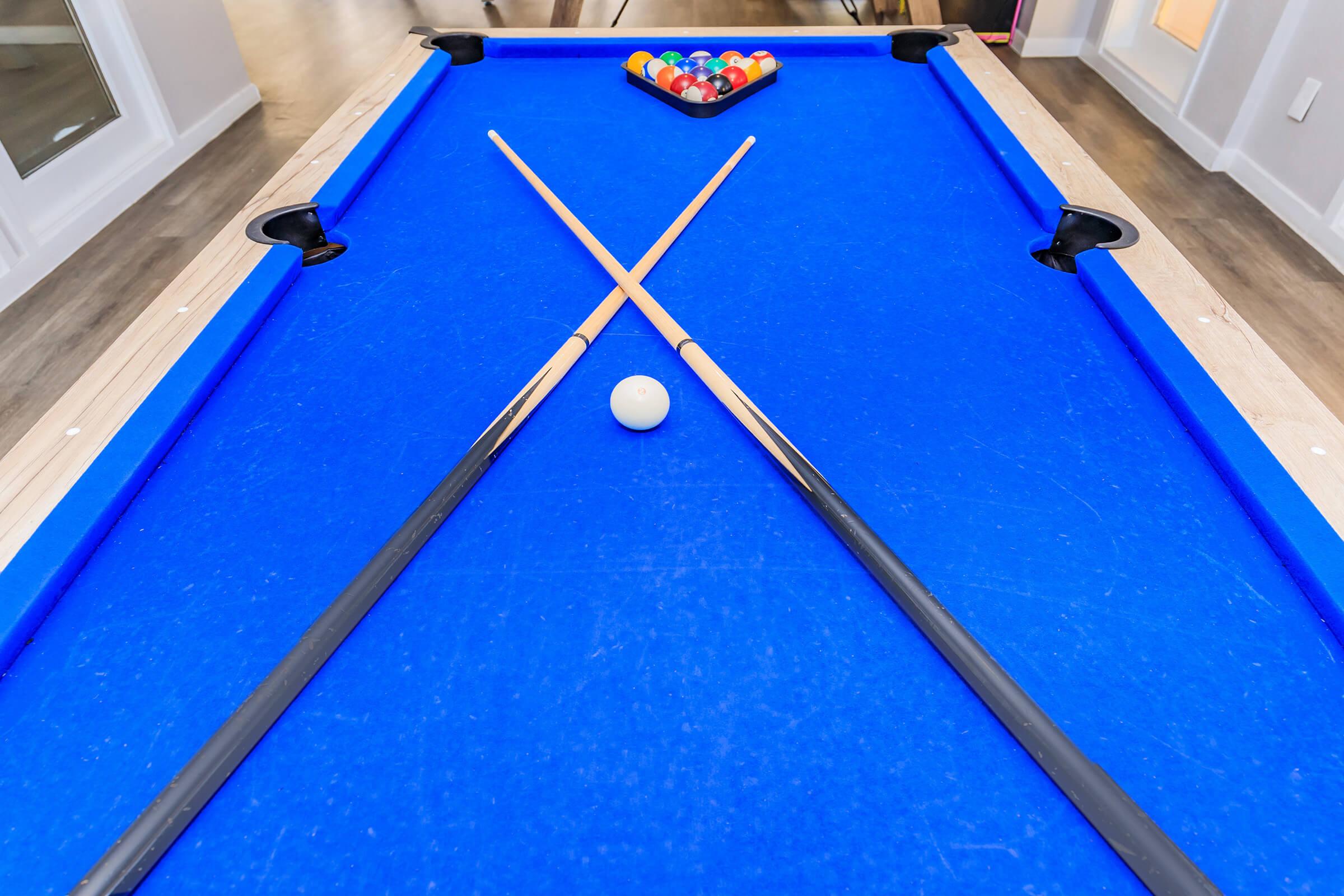A blue pool table featuring crossed cue sticks and a cue ball positioned in the center. At the far end, a triangle rack holds a set of colorful billiard balls, ready for a game. The setting is bright and modern, with light-colored flooring and walls.