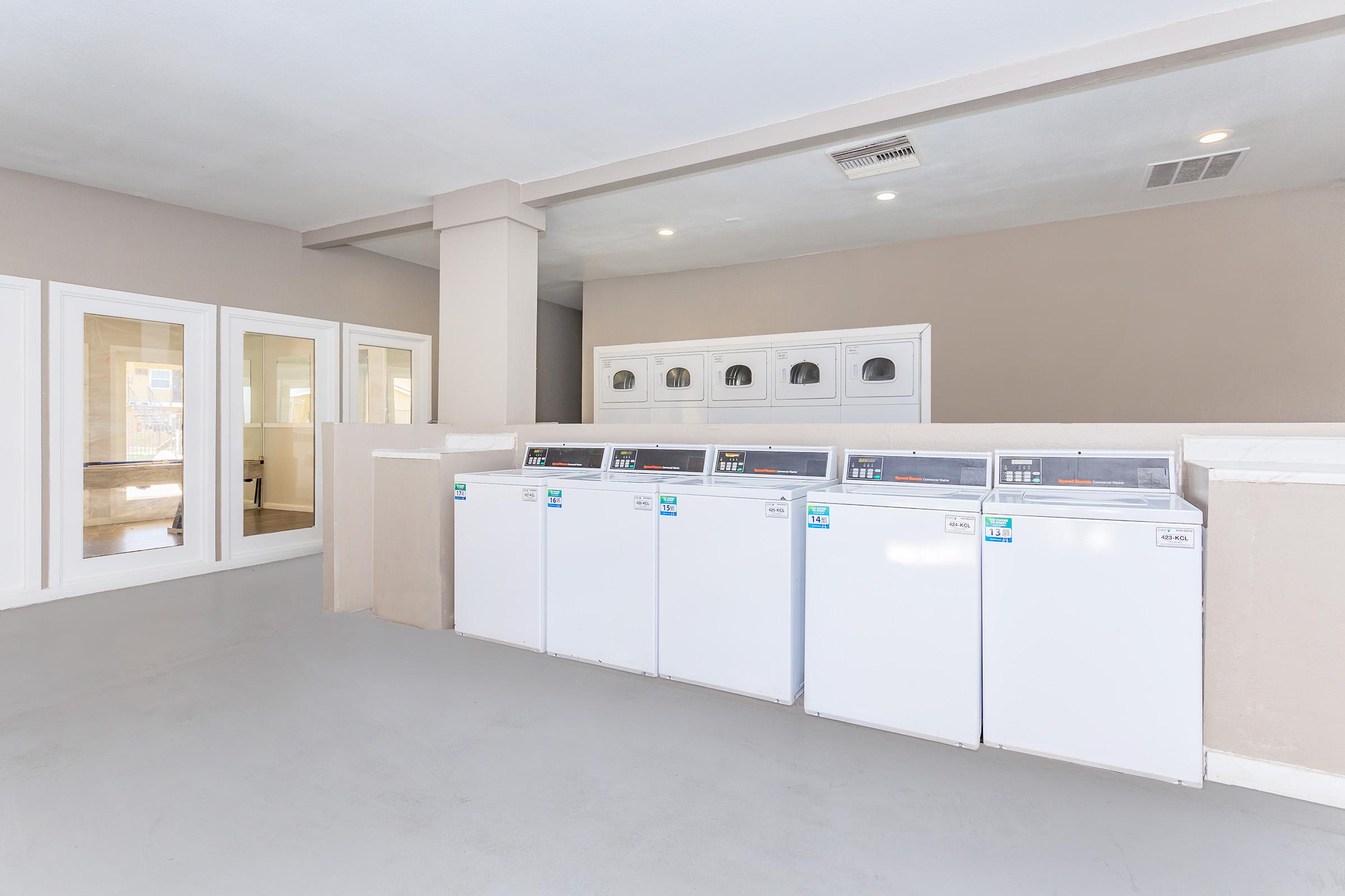 A clean laundry room featuring several white washing machines lined up against a beige wall. The room has a minimalist design with light gray flooring and large windows allowing natural light. There are also doorways leading to other areas, creating an open and spacious feel.