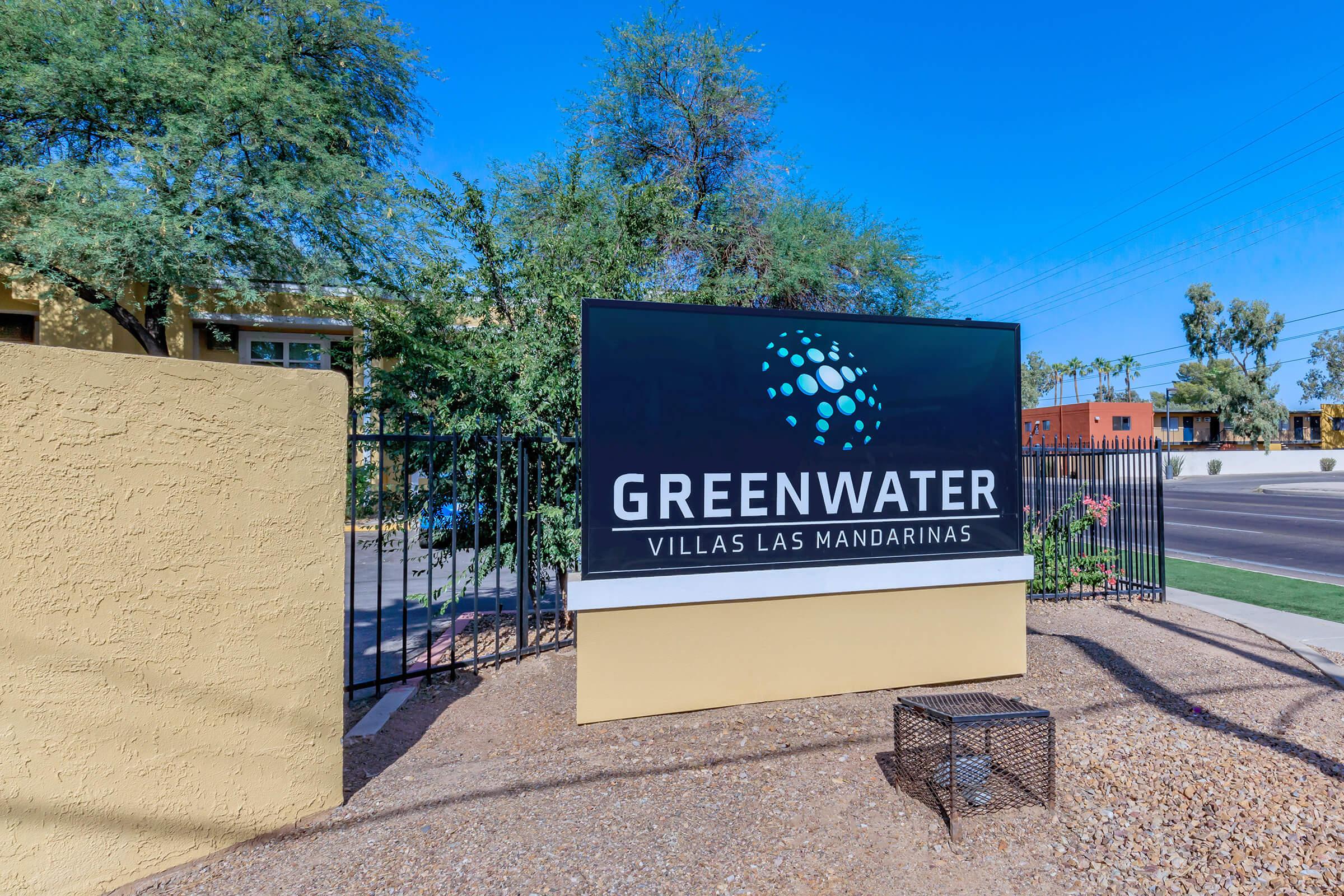 Sign for Greenwater Villas Las Mandarinas, featuring a modern design with blue and green graphics. The sign is situated outside, with a desert landscape in the background, including sparse vegetation and a clear blue sky. A fenced area surrounds the sign, indicating an apartment or residential community.