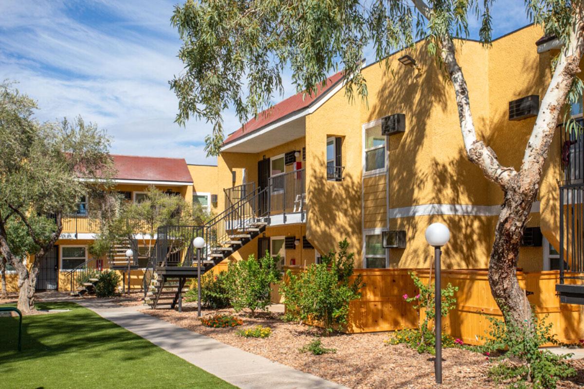 A sunny view of a multi-unit apartment complex featuring yellow exterior walls, with landscaped gardens, pathways, and a few trees. The buildings have staircases leading to the upper floors, and outdoor lighting fixtures enhance the inviting atmosphere. Blue sky and wispy clouds are visible above.