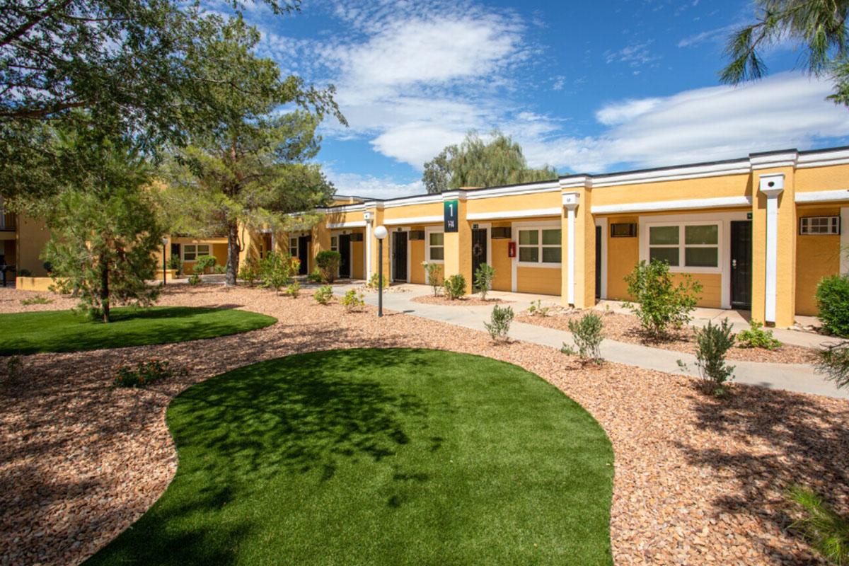 Landscape view of a colorful building with entrances along a pathway. Lush green grass and carefully arranged shrubs surround the structure, with a blue sky and scattered clouds above. The scene conveys a welcoming and well-maintained atmosphere.
