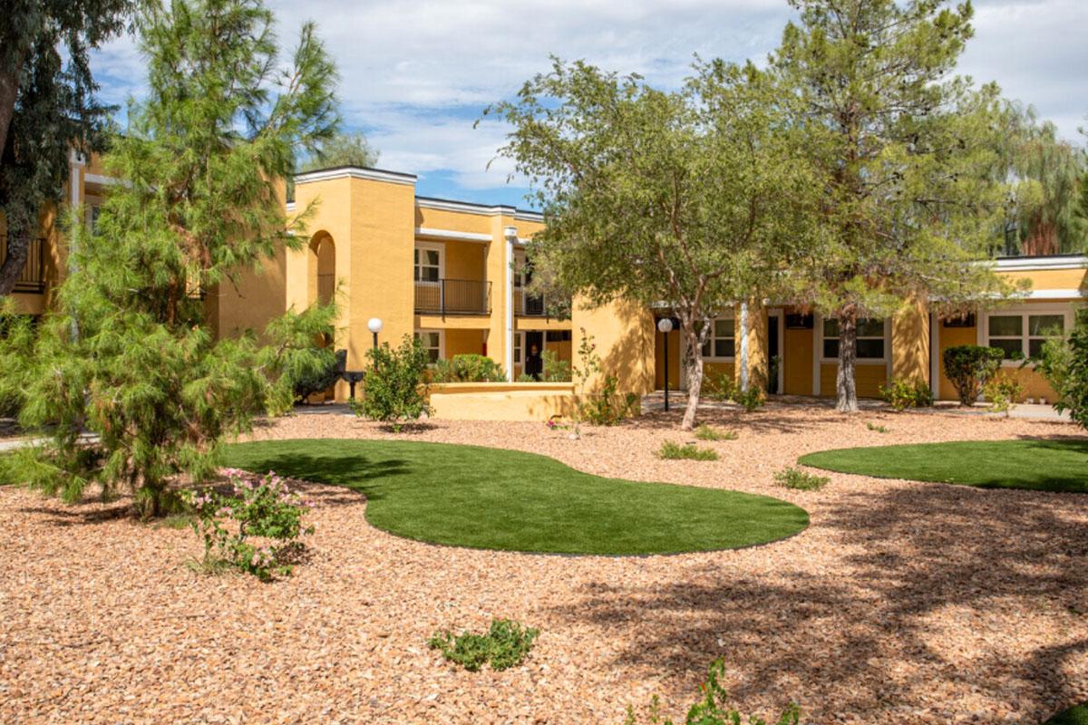 A well-maintained outdoor area featuring a yellow building with multiple units, surrounded by lush green grass and trees. The landscape includes gravel paths and landscaped shrubs, creating a serene environment ideal for relaxation. The sky is partly cloudy, adding to the peaceful atmosphere.