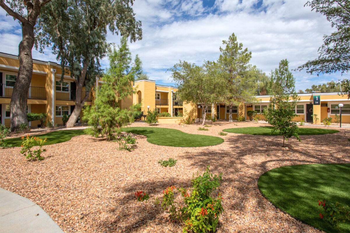 A landscaped courtyard featuring bright green grass and small trees, surrounded by buildings painted in warm yellow tones. The area is dotted with decorative rocks and shrubs, creating a welcoming outdoor space under a partly cloudy sky.