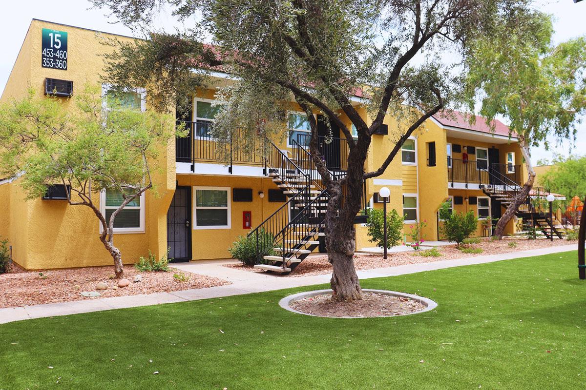 Two-story apartment building painted yellow, featuring multiple units with balconies. The building has black metal stairs leading to the second floor. Surrounding the structure is a green lawn with desert landscaping, including small trees and light fixtures. The area is bright and inviting.