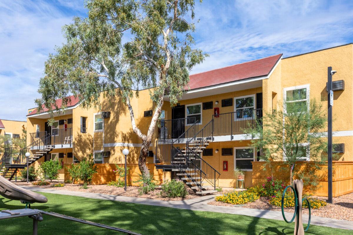 A sunny outdoor view of a two-story apartment complex with yellow walls, featuring green landscaping, trees, and playground equipment in the foreground. There are stairs leading to the second floor, and the setting has a clear blue sky with a few clouds.