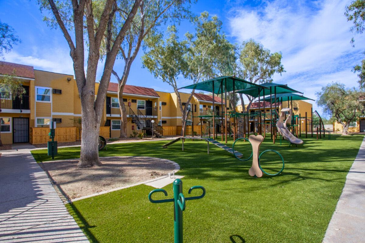 A landscaped playground area in front of yellow apartment buildings, featuring a green turf lawn, climbing structures, a slide, and a large dog bone sculpture. Surrounded by trees, the space is designed for children and pets. Clear blue skies overhead enhance the cheerful atmosphere.