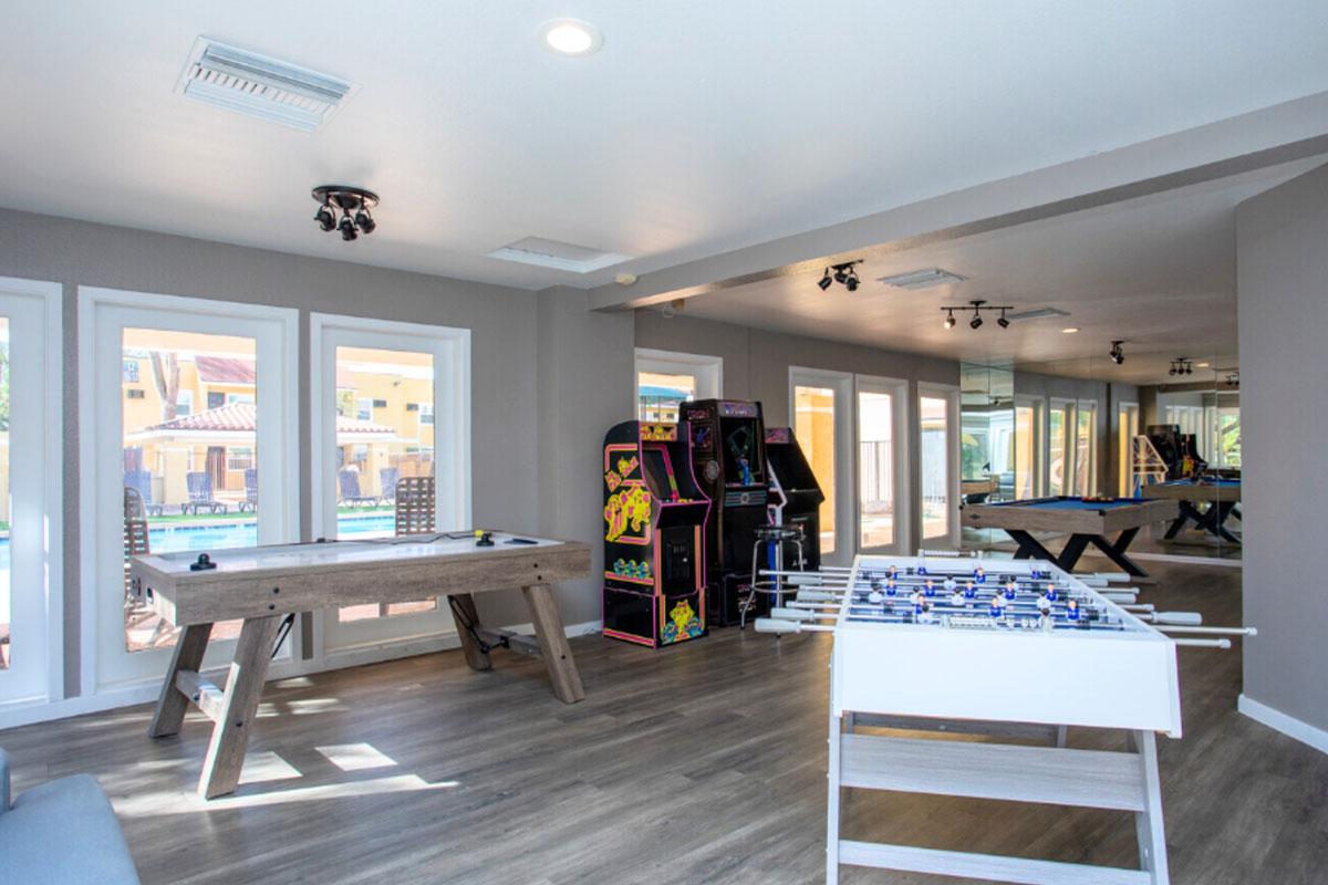 Interior view of a recreational room featuring a foosball table, a pool table, and classic arcade games. Large windows allow natural light, and there’s an outdoor pool visible through the glass doors. The space has modern decor with wooden flooring and a relaxed atmosphere.