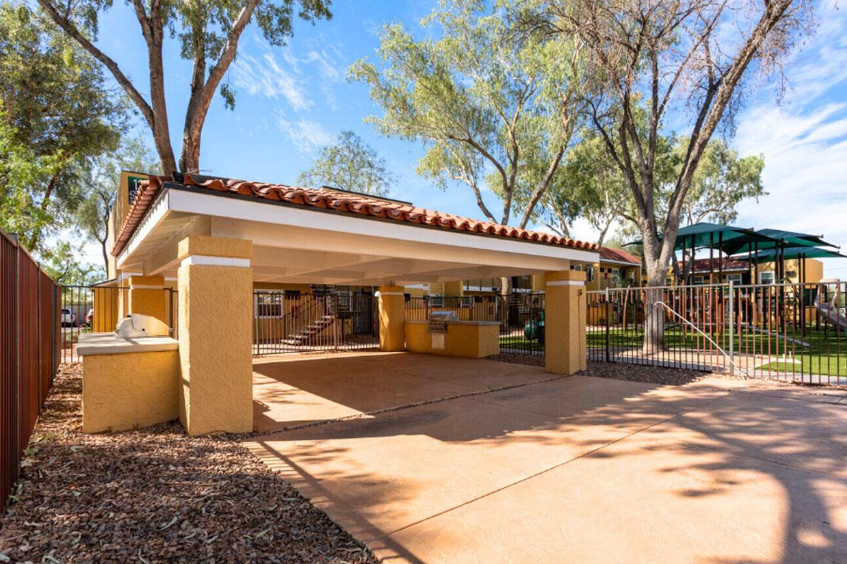 A covered outdoor space with a stucco exterior and a terracotta tile roof, surrounded by trees. There are shaded areas with tables and chairs visible in the background, along with a fenced area and playground equipment nearby, all under a clear blue sky.