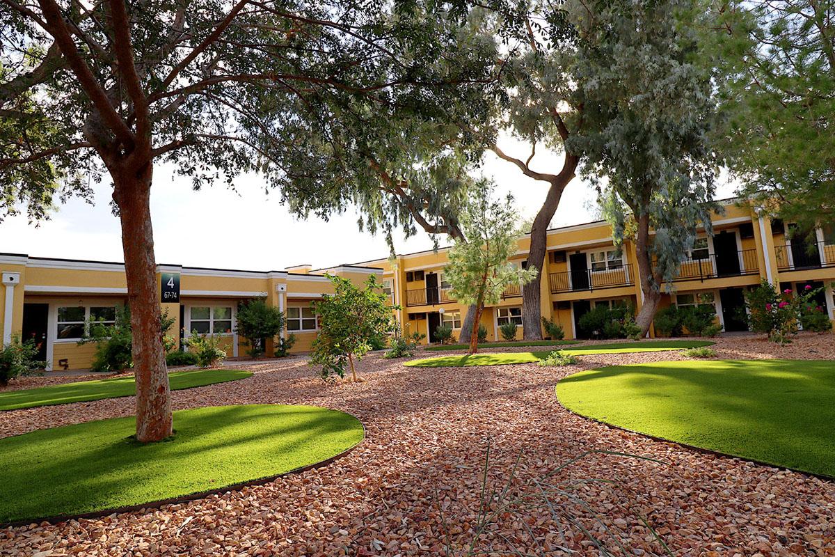 A landscaped courtyard featuring lush green grass areas, surrounded by decorative gravel. Several trees provide shade, and a building with yellow walls and black accents is visible in the background. The setting appears tranquil and well-maintained, suitable for relaxation.