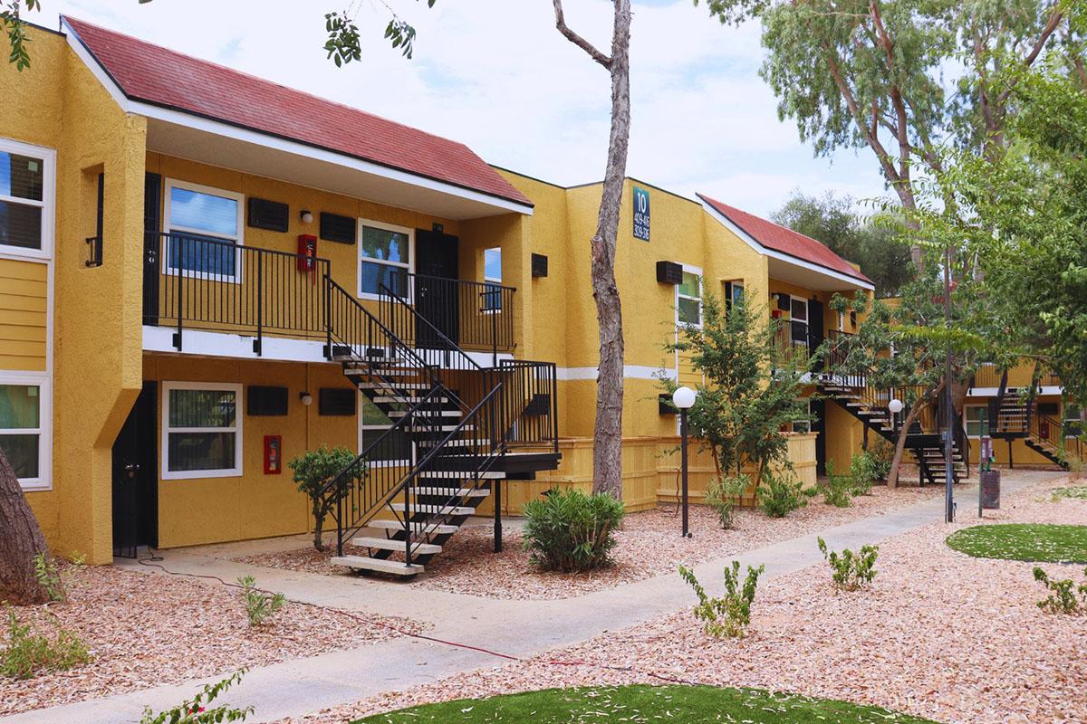 A view of a yellow apartment complex with two-story buildings featuring balconies. Pathways and small landscaped areas with shrubs and rocks are visible. Trees provide shade, and an outdoor light fixture stands near the walkway. The overall atmosphere is inviting and residential.