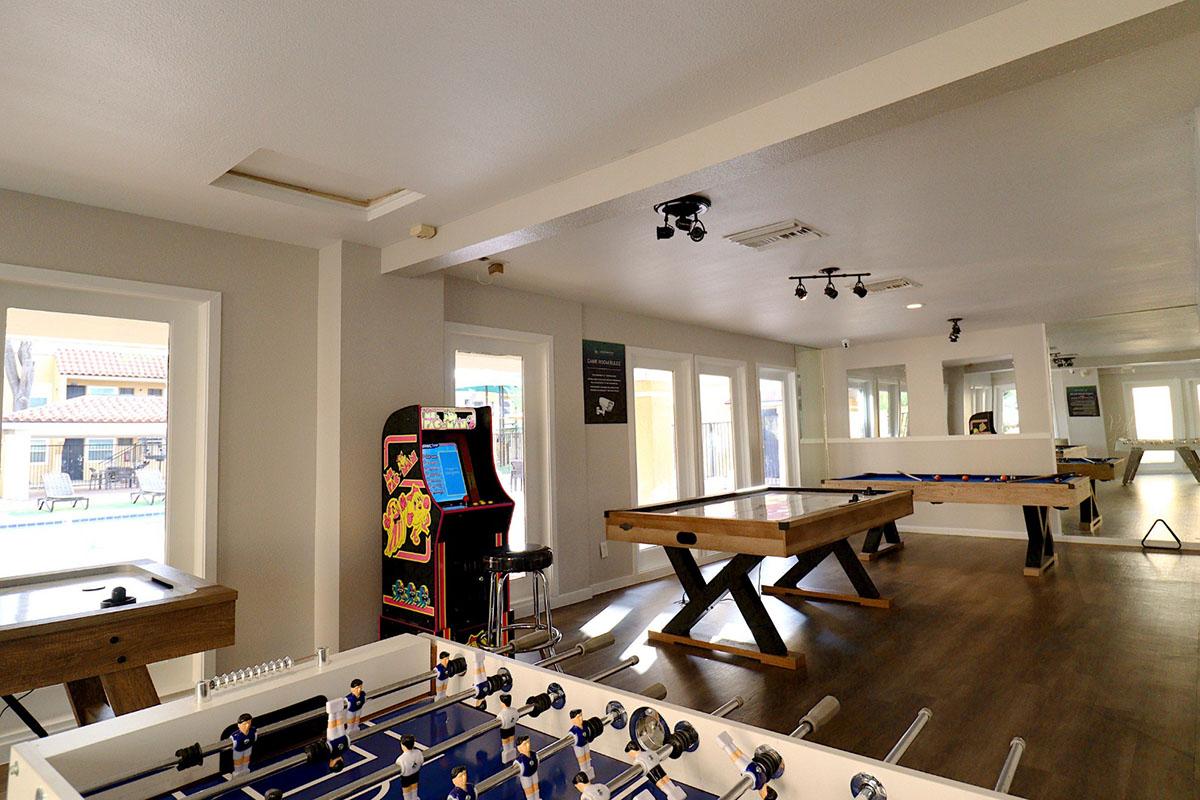 A spacious game room featuring a foosball table, a pool table, and an arcade machine. Large windows allow natural light to fill the space, with modern decor and hardwood flooring. The area is designed for recreation and entertainment, providing a casual atmosphere for socializing and playing games.