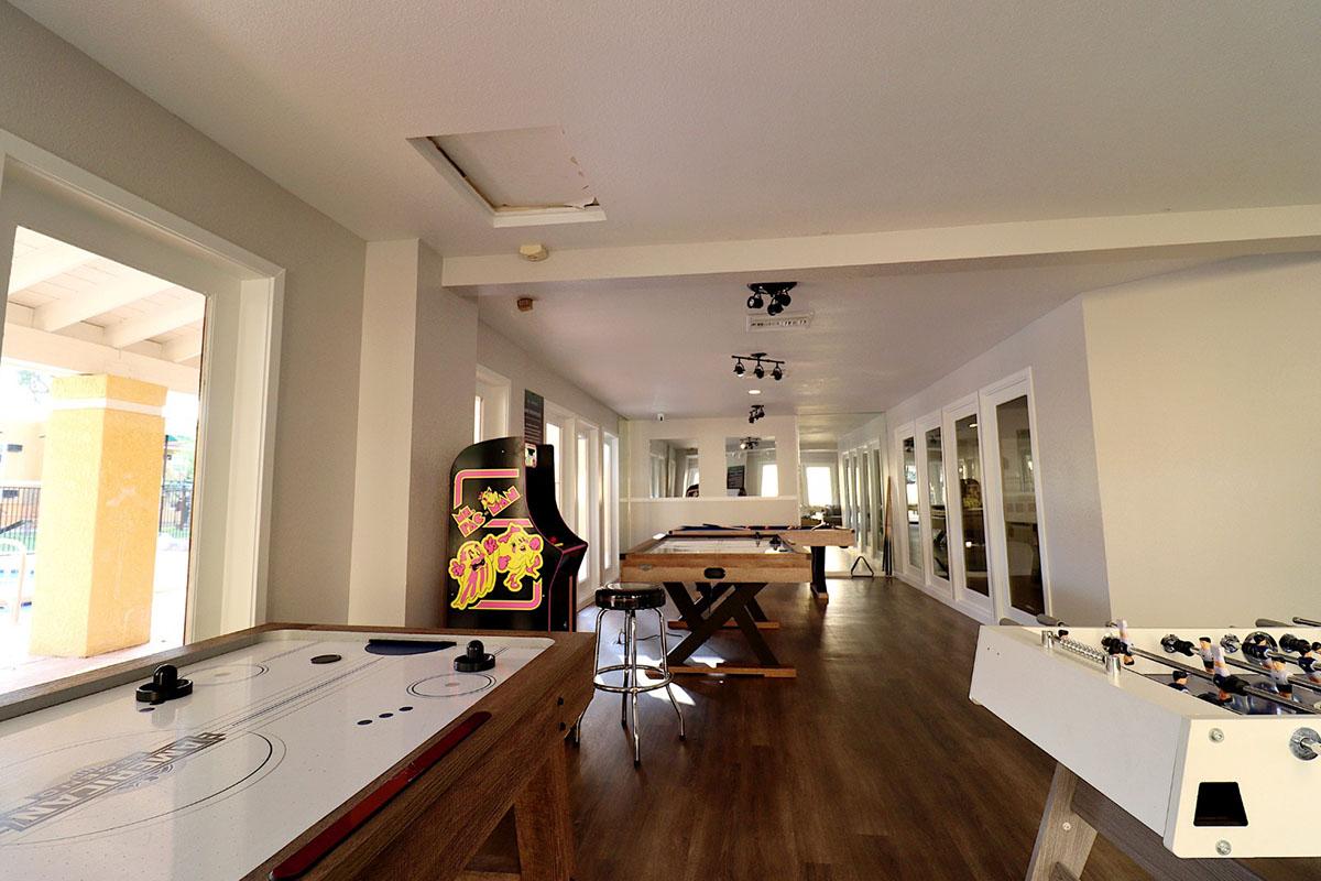 A spacious game room featuring an air hockey table, foosball table, and several tables in a well-lit environment. Large windows allow natural light to fill the room, creating a welcoming atmosphere for leisure and social activities.