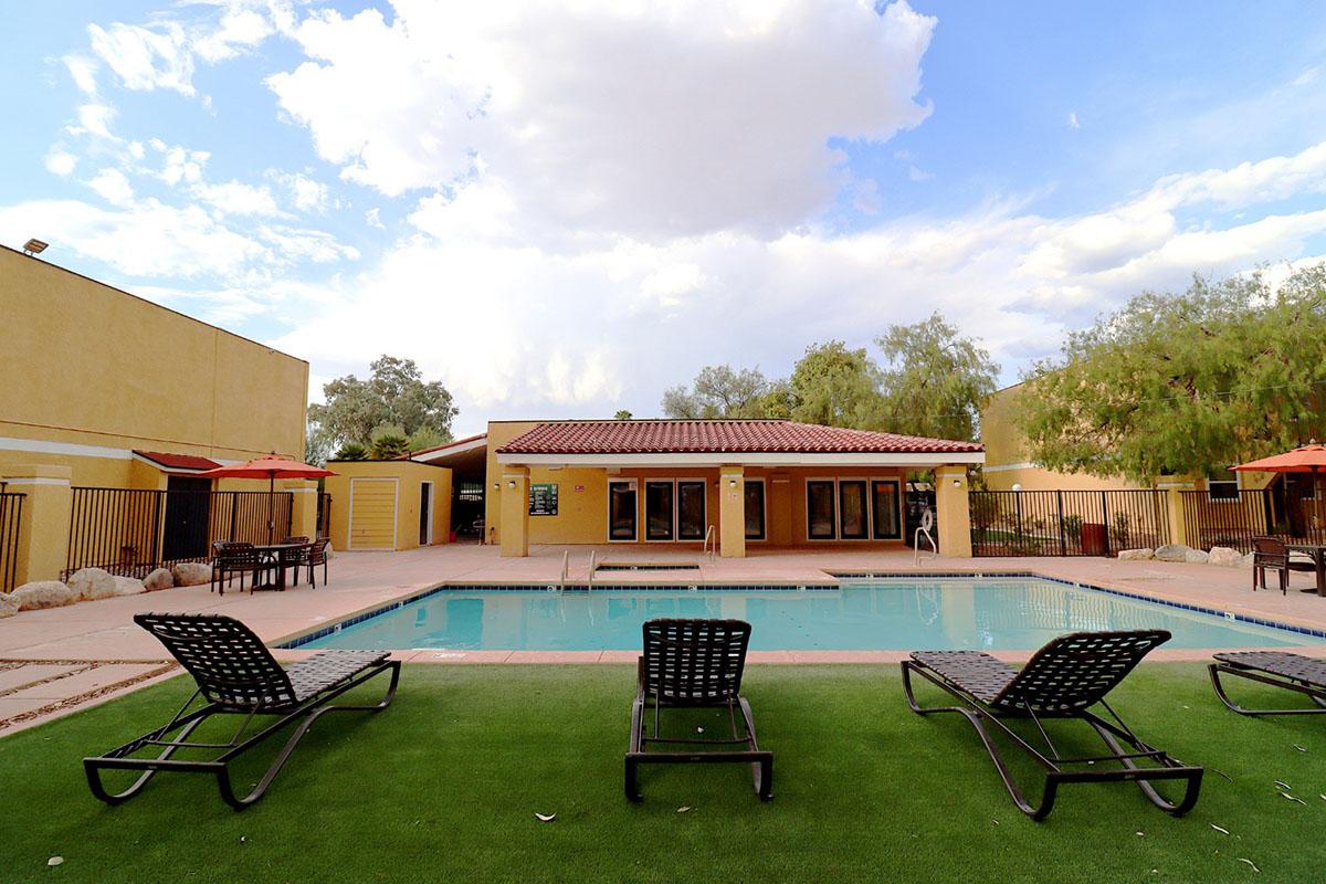 A swimming pool surrounded by lounge chairs on a grassy area. In the background, there is a yellow building with several windows and a roofed outdoor space. The sky is partly cloudy, and there are a few trees nearby, adding to the serene atmosphere.