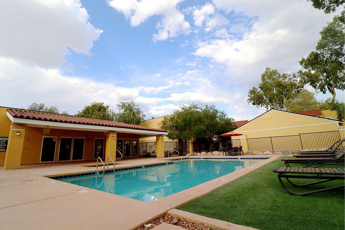 A serene outdoor pool area featuring a clear blue pool surrounded by lounge chairs. The pool is bordered by a small grassy area and a few trees, with a backdrop of sunny skies and scattered clouds. Nearby, there are building structures with a warm color palette, enhancing the inviting atmosphere.