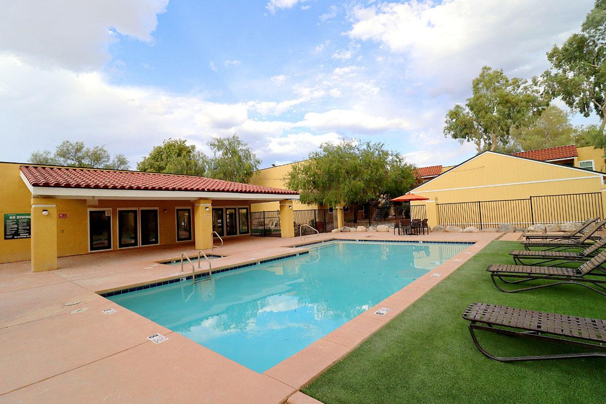 A swimming pool surrounded by lounge chairs, set against a backdrop of yellow buildings and green trees. The pool area features a clean, inviting atmosphere with a clear sky above and well-maintained landscaping, perfect for relaxation and leisure.