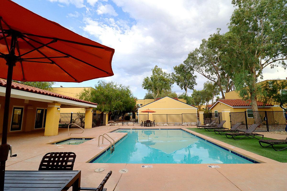 A sunny outdoor swimming pool area with a turquoise pool surrounded by lounge chairs and umbrellas. In the background, there are yellow buildings and trees. The scene is tranquil, with a few clouds in the sky, creating a relaxing atmosphere.