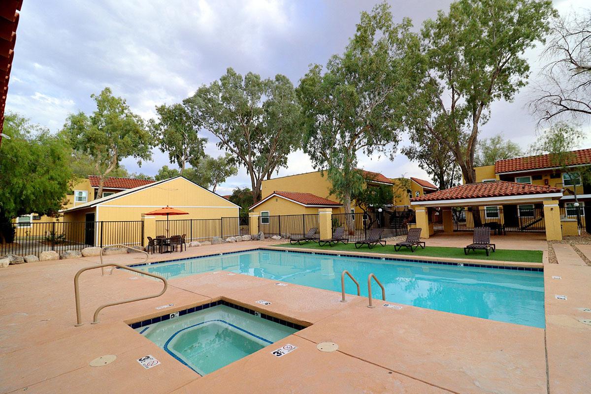 A swimming pool surrounded by trees and lounge chairs, with a small hot tub nearby. There are yellow buildings in the background, and an umbrella at a seating area. The sky is overcast, creating a calm atmosphere.