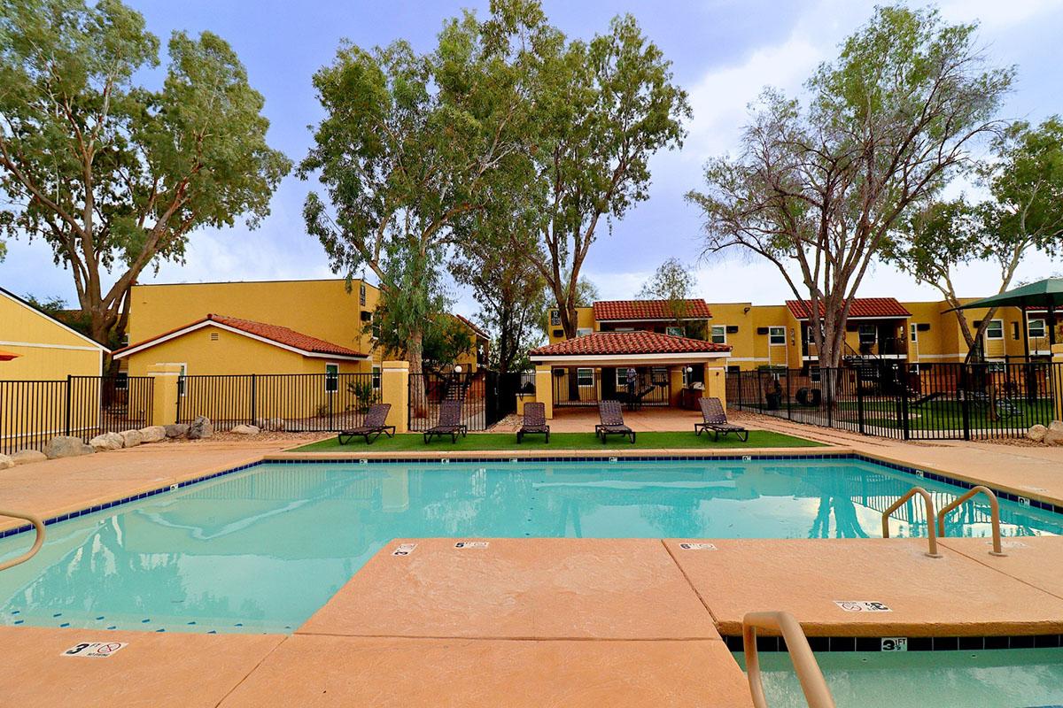 A sunny outdoor swimming pool surrounded by lounge chairs and tall trees. In the background, colorful buildings with yellow facades and red rooftops are visible. The pool area is fenced, creating a relaxing atmosphere ideal for leisure and recreation.
