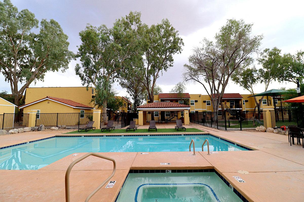 A swimming pool area with a hot tub, surrounded by trees and lounge chairs. Nearby, there are yellow buildings with a Spanish-style roof, a patio, and outdoor seating. The sky is partly cloudy, creating a serene, inviting atmosphere for relaxation.