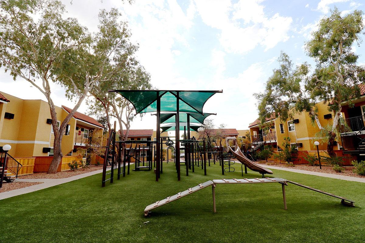 A playground area featuring green turf, a climbing structure with slides, and shaded areas underneath large canopies. Surrounding the playground are residential buildings with a bright color scheme and trees, set against a partly cloudy sky.