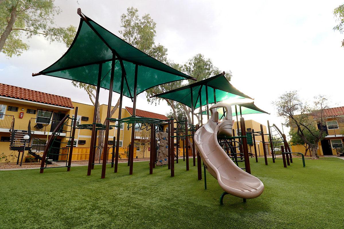 Playground with a large, twisting slide, shaded by green fabric canopies. The area is surrounded by grassy turf and features climbing structures. Yellow apartment buildings are visible in the background, creating a vibrant outdoor play space.