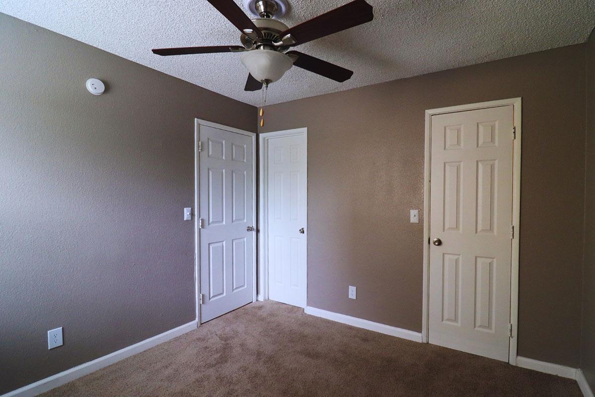 A small, neutral-colored room featuring a ceiling fan, two closed doors—one white and one beige—and beige carpet. The walls are painted a light brown hue, and there is a small white light switch on the left wall.