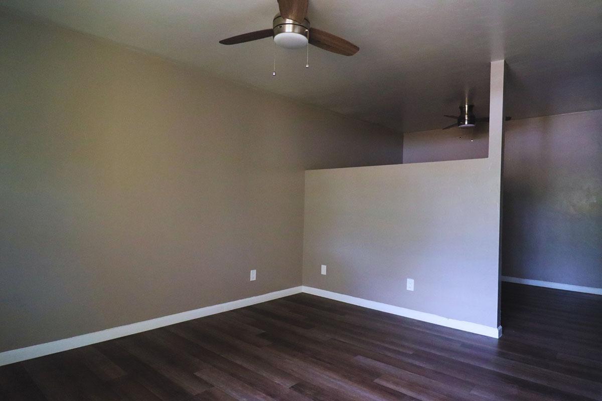 An empty room with light brown walls and laminate flooring. A ceiling fan is mounted in the center, and there is a partial wall separating the space from another area. Natural light may be entering from an unseen source. The overall atmosphere is minimalistic and unadorned.