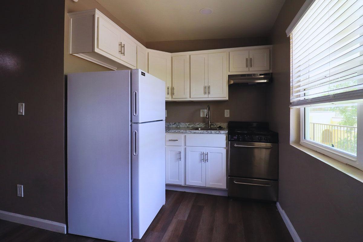 A modern kitchen featuring white cabinets, a stainless steel stove, and a large refrigerator. The countertop is decorated with a light-colored granite, and there is a window providing natural light. The walls are painted a soft brown, and the flooring is dark wood.