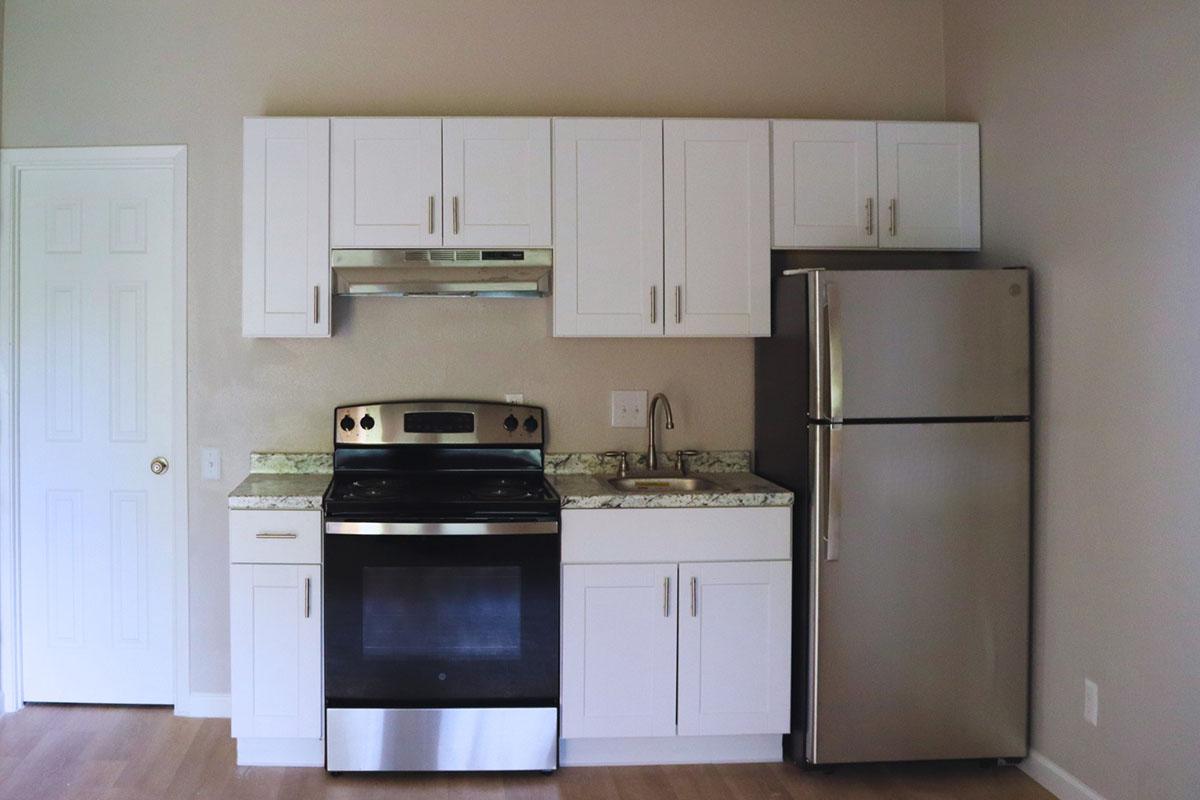 A modern kitchen featuring white cabinetry, a stainless steel stove with an oven, a matching microwave, and a refrigerator. The countertops are made of light-colored granite. The space has beige walls and wooden flooring, creating a bright and clean aesthetic.