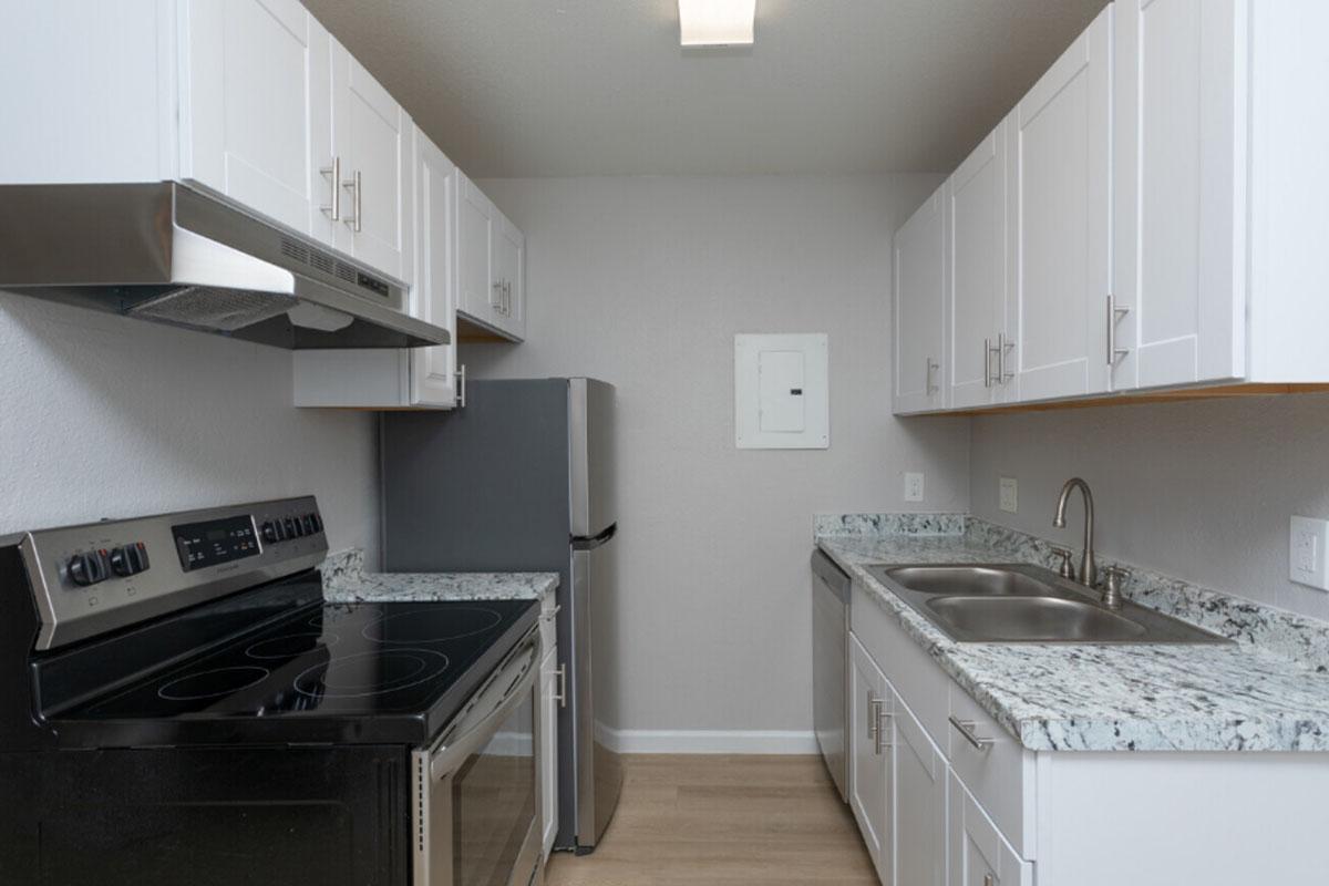 Modern kitchen featuring white cabinets, stainless steel appliances including an oven and refrigerator, dual sink with a chrome faucet, and a granite countertop. The walls are painted in a light color, and the flooring is wood-like laminate. Bright lighting is present overhead.