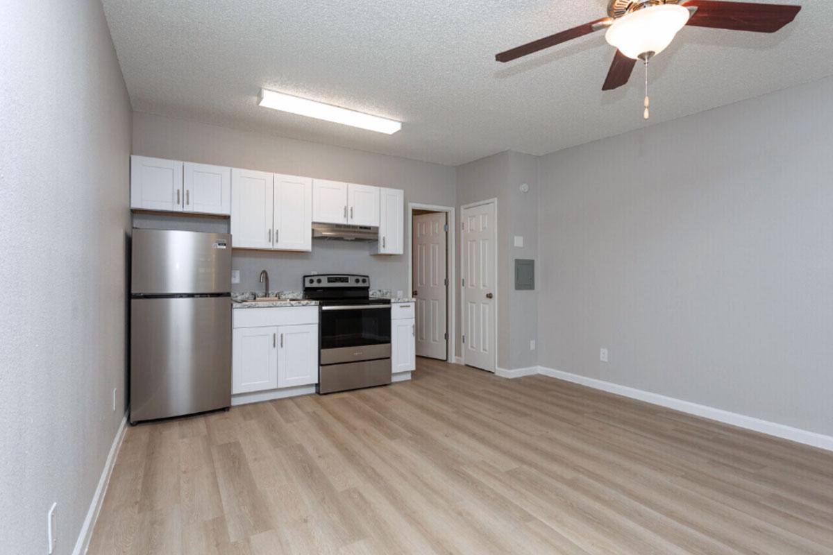 A modern kitchen and living area featuring light wood flooring, white cabinetry, stainless steel appliances including a refrigerator and stove, and a ceiling fan. The walls are painted in a light gray color, creating a bright and inviting atmosphere.