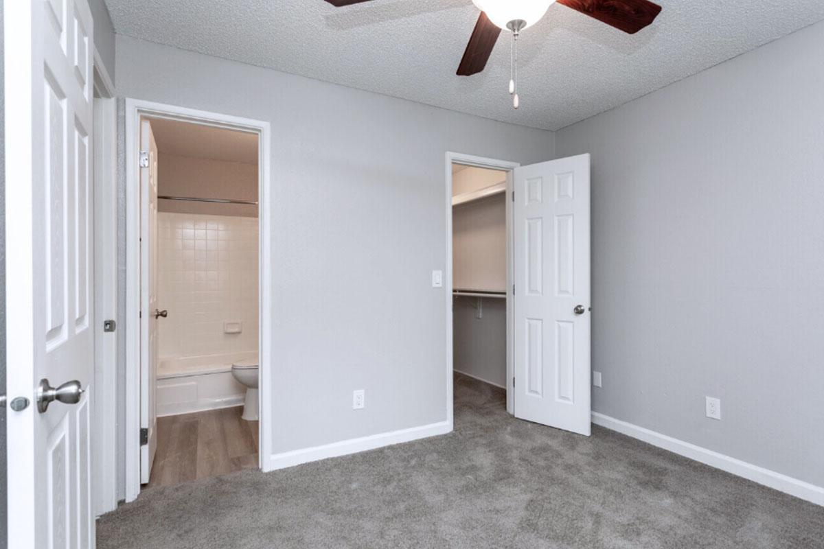 A well-lit room featuring light gray walls and carpet. There is a ceiling fan with wooden blades. Two doors are visible: one leads to a bathroom with a bathtub, and the other opens to a closet. The room has a minimalistic design, creating a spacious and inviting atmosphere.