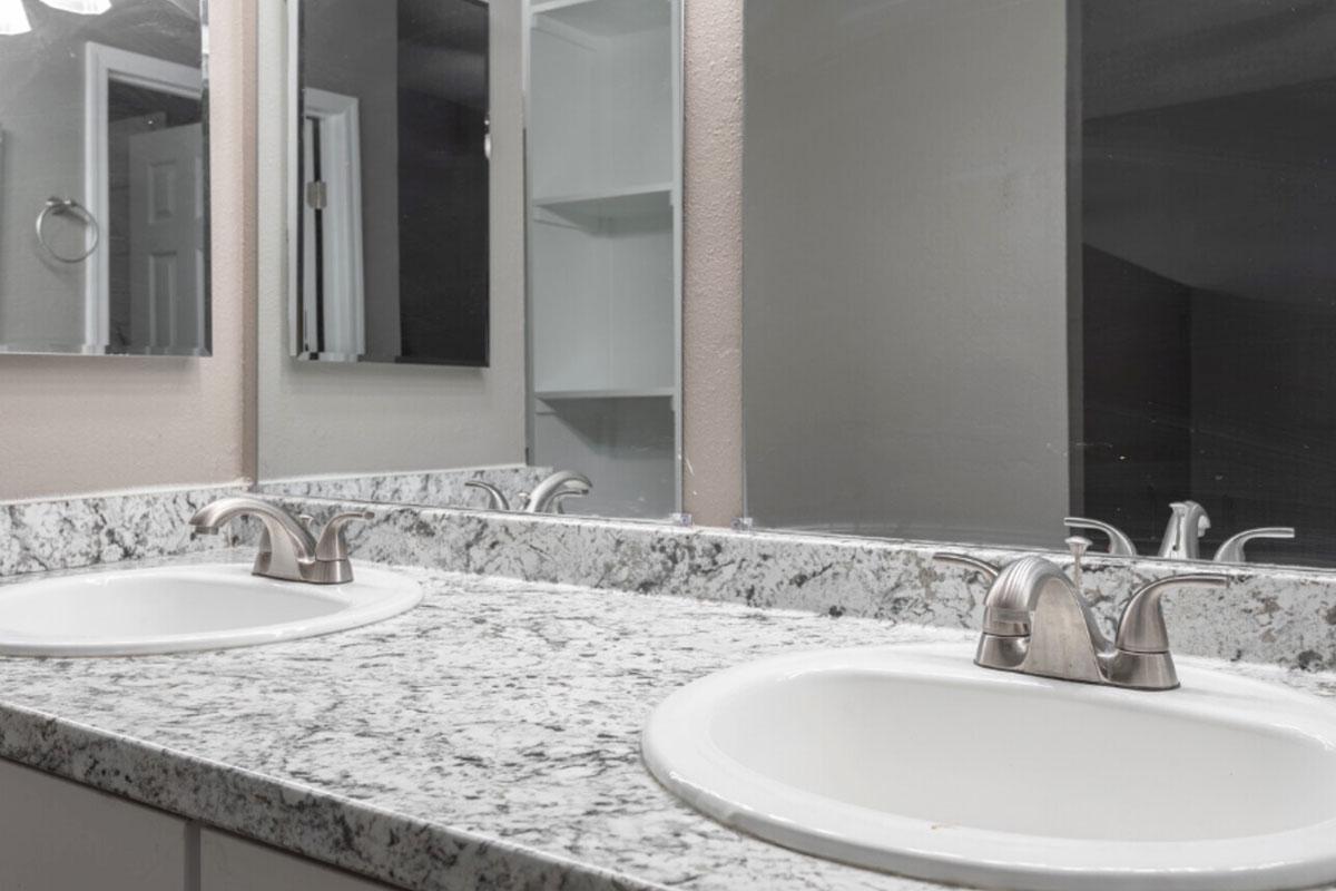 Dual sinks in a modern bathroom vanity with a light-colored granite countertop. A large mirror above the sinks reflects the space. The walls are painted a soft color, and there is a shelf to the side. Overall, the design is clean and contemporary.